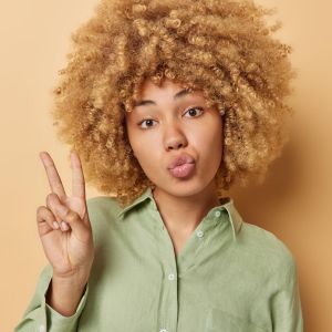 pretty-young-european-woman-with-curly-bushy-hair-keeps-lips-folded-shows-victory-v-sign-wears-green-shirt-isolated-brown-studio-background-celebrates-win-people-body-language-concept_273609-61412