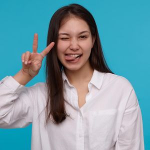 studio-portrait-young-funny-brunette-female-with-long-straight-hair-showing-her-tongue-v-sign-wink-isolated-blue-background_295783-16248
